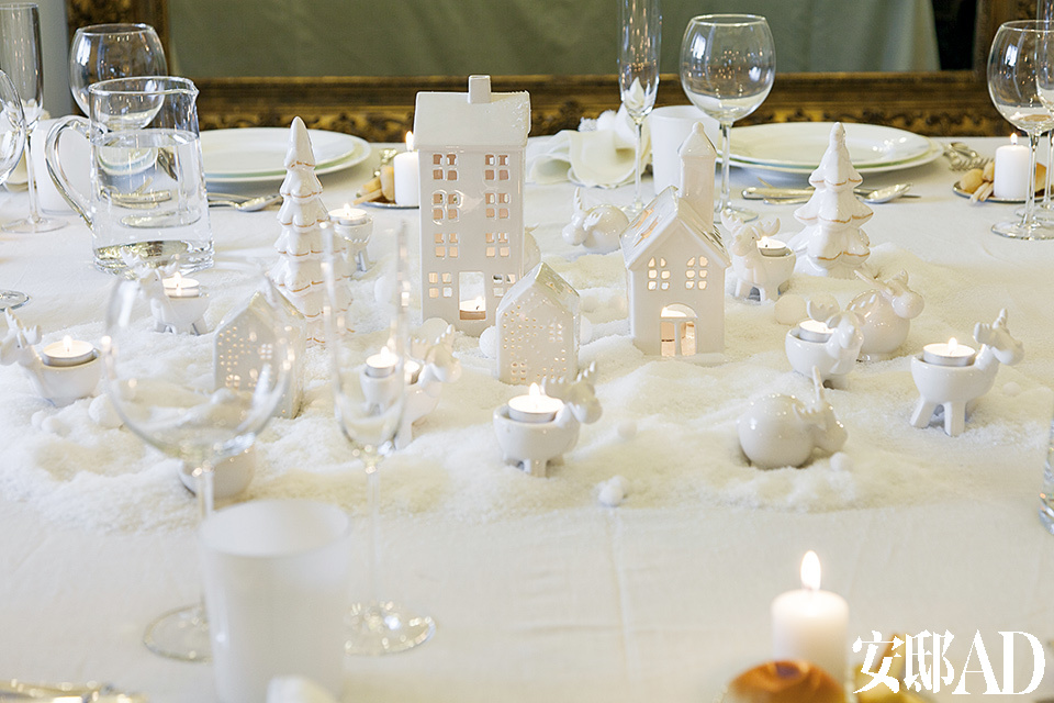 整个圣诞餐桌的布置也是纯白的主题，带有童话色彩的小屋和着点点烛光烘托出家的温暖。圣诞餐桌的布置细节：百分百纯麻质地的桌布是定制产品。瑞典风格的白色瓷蜡烛屋显现出童话般的纯真，烛光照着白色的瓷质麋鹿，营造出特殊的光影，为整个房间增添了一丝梦幻的魔术色彩。白色骨瓷餐盘和水晶杯都是设计师精心挑选的。