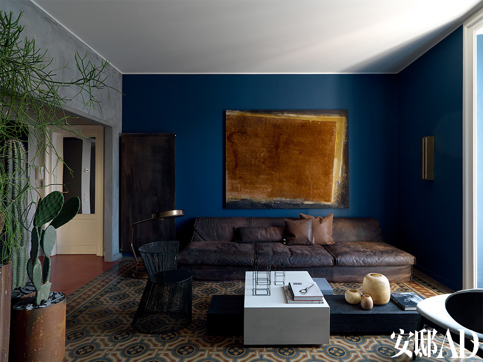 老旧的地砖，以质朴、静默的色调，主导着客厅。宽大的棕色皮质沙发占据了几乎一整侧的位置，沙发上方深蓝色墙面上悬挂着的艺术品，与一旁硕大的金属落地阅读灯，都使用了相近颜色。暖气被藏在一块巨大的金属板后面，与室内陈设所使用的基调和谐地相处。
